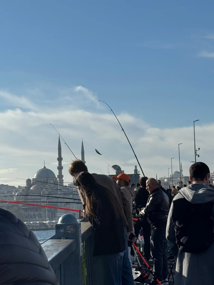 People fishing from the Galata Bridge, a fish flying up on a line, with a mosque behind