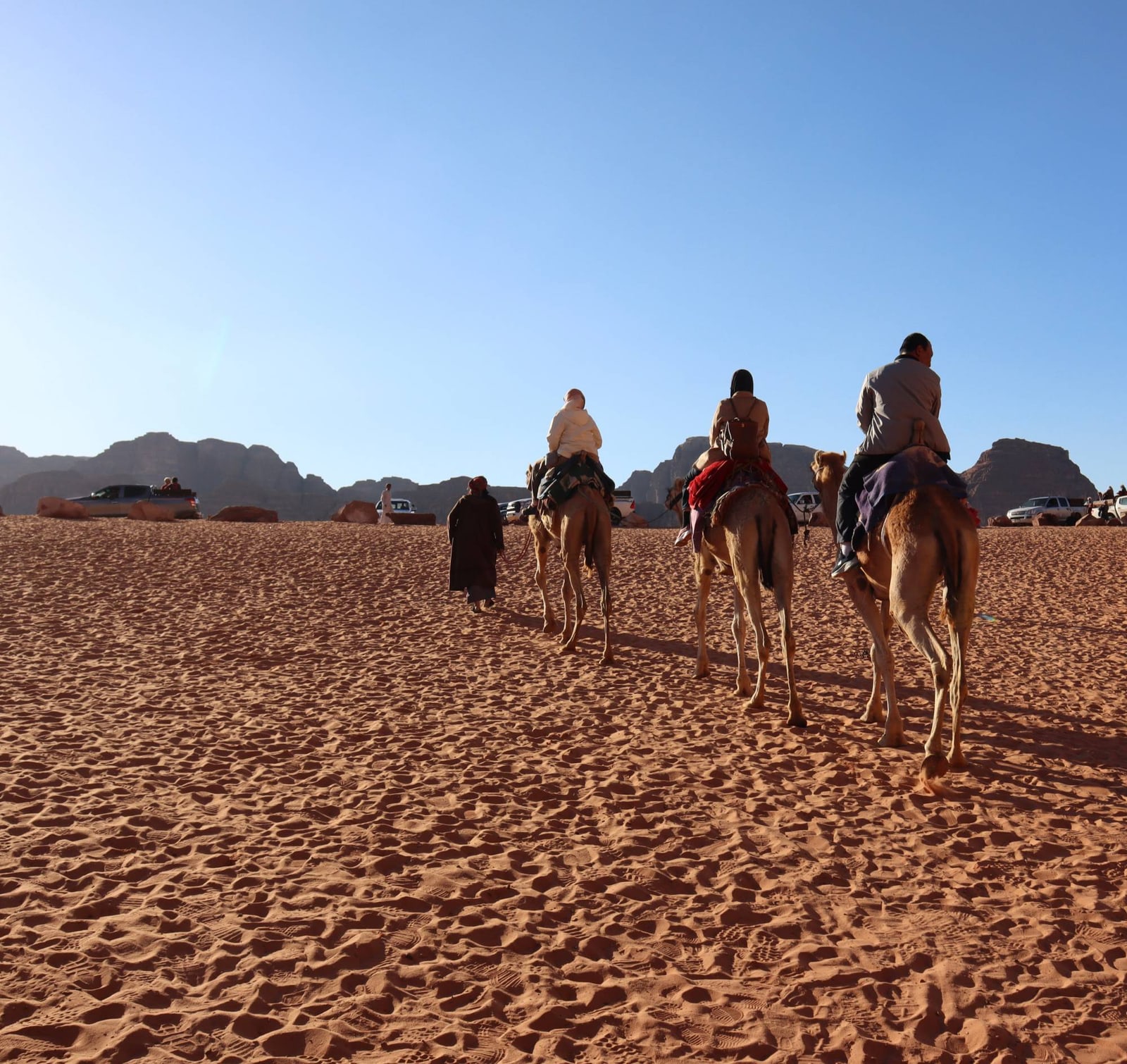 Three people riding camels across red desert sand towards rocky mountains under a clear blue sky, led by a guide
