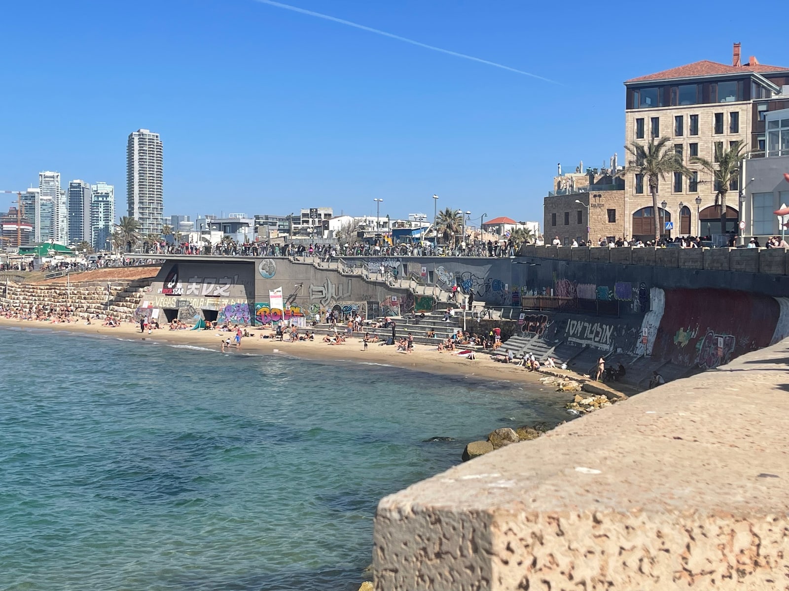 A sunny seafront: turquoise water and a sandy beach below a stone sea wall, with city towers in the distance